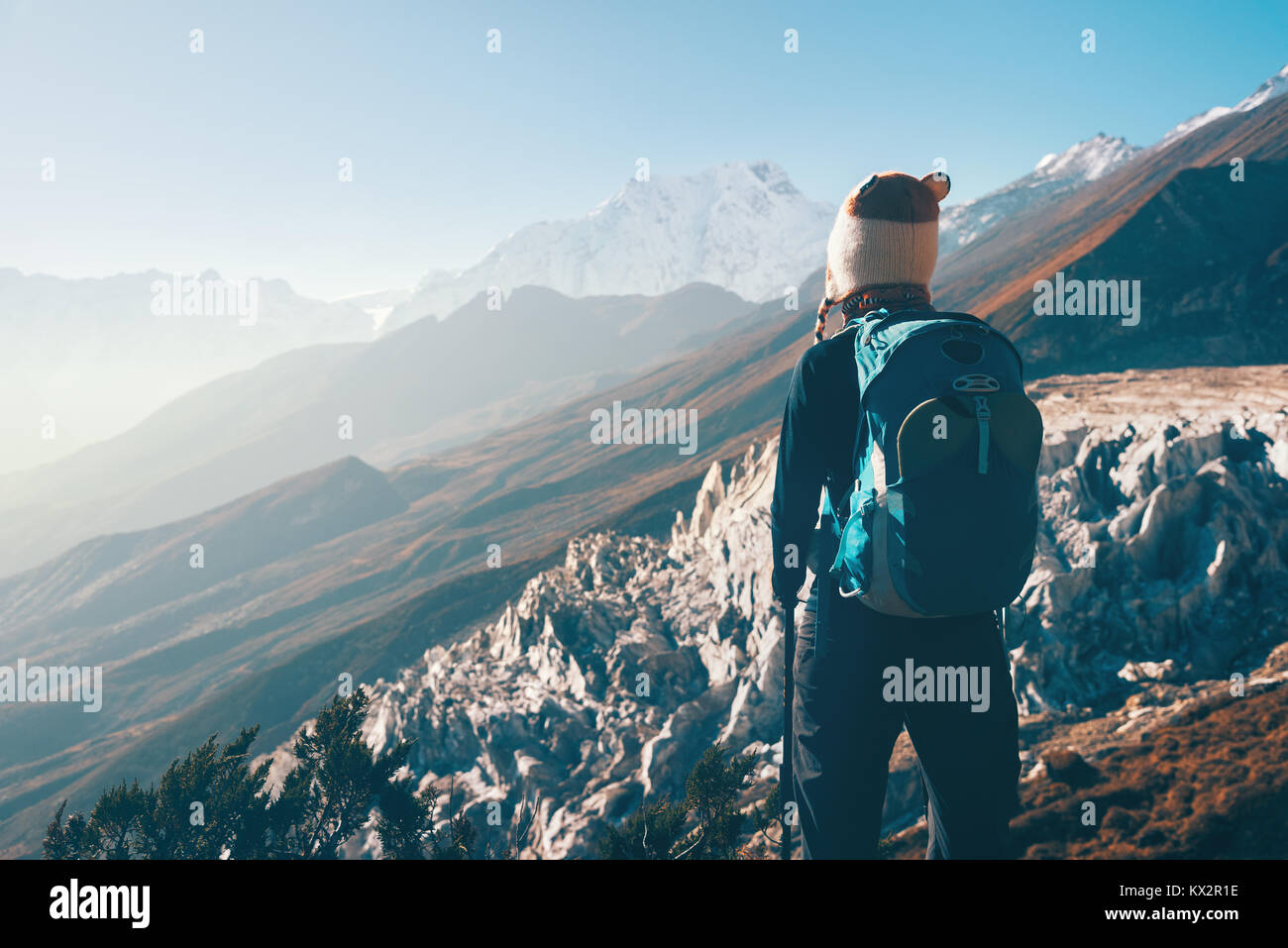 Standing young woman with backpack on the mountain peak and looking on beautiful mountain valley at sunset. Landscape with girl, rocks with snowy peak Stock Photo