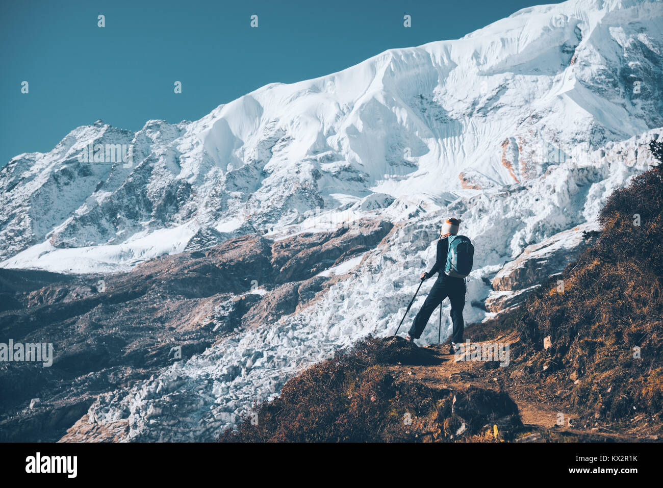 Standing young woman with backpack on the mountain peak and looking on beautiful mountains and glacier at sunset. Landscape with girl, rocks with snow Stock Photo