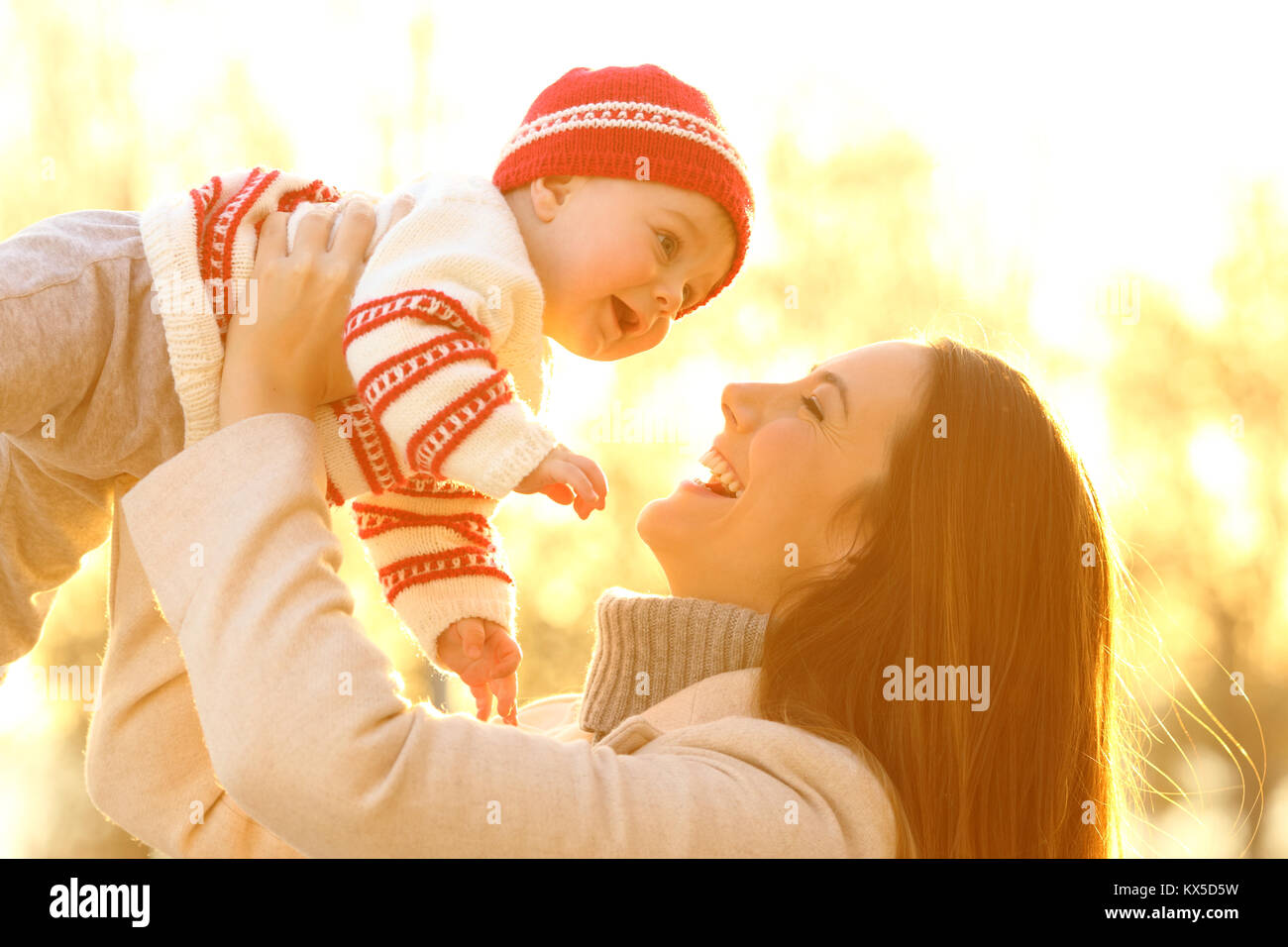Backlighting portrait of a joyful mother raising her baby outdoors at sunset in winter Stock Photo