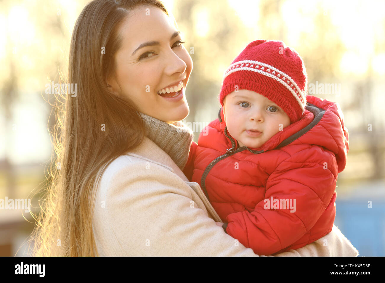 Portrait of a smiley mother posing with her baby keeping warm wearing a red jacket outdoors in winter Stock Photo