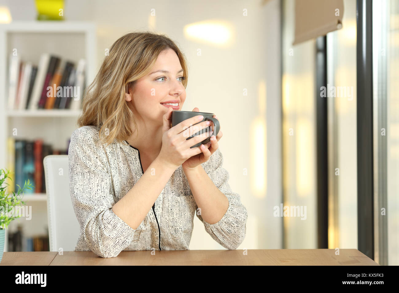 Portrait of a woman looking through a window drinking coffee sitting in a table at home Stock Photo