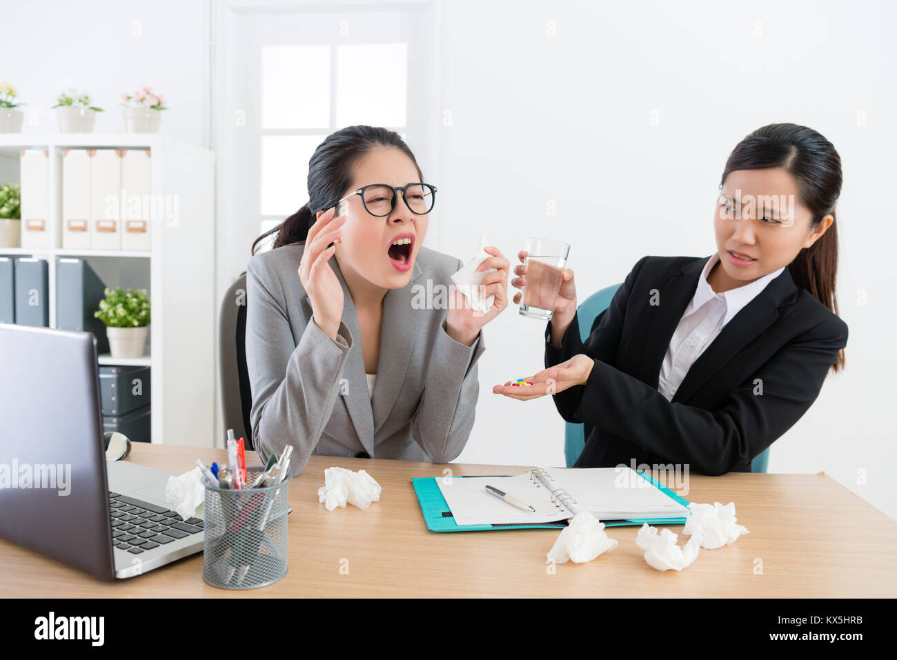 business woman sneezing while her worker partner looking at her feeling fear in office and giving pills with water. Stock Photo