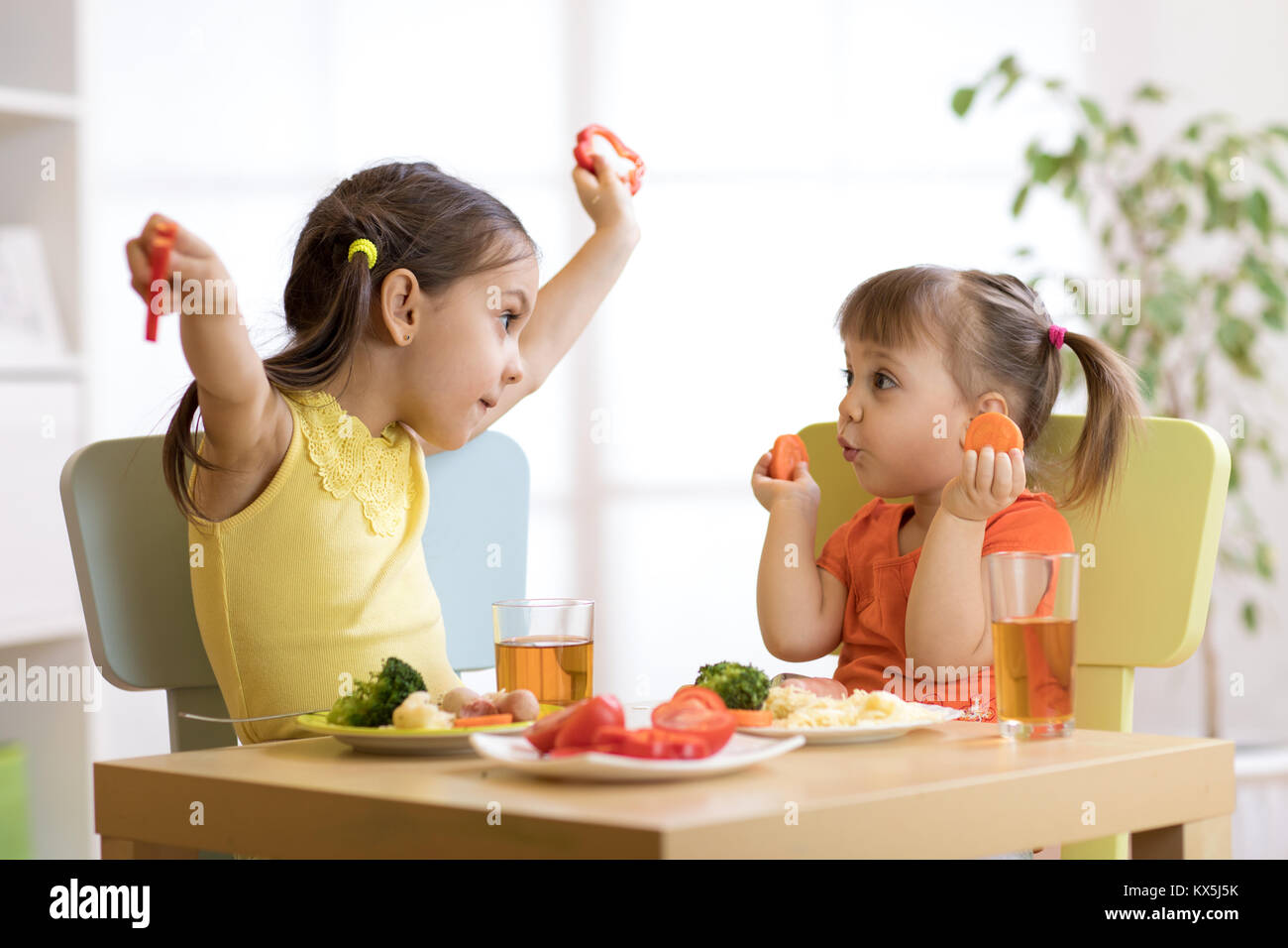 Cute smiling child and kid toddler girls playing and eating spaghetti with vegetables for healthy lunch sitting in a white sunny kitchen with big wind Stock Photo