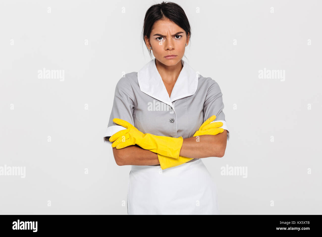 Portrait of grumpy attractve maid in uniform and yellow rubber gloves standing with crossed hands and looking at camera, isolated on white background Stock Photo