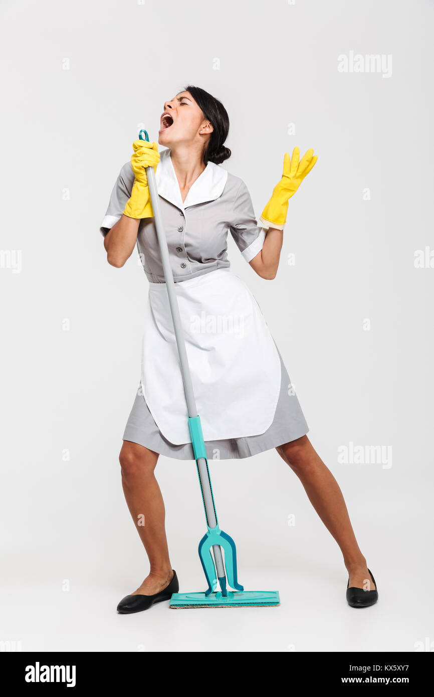 Full length portrait of funny housekeeper in uniform singing in mop as microphone, isolated on white background Stock Photo