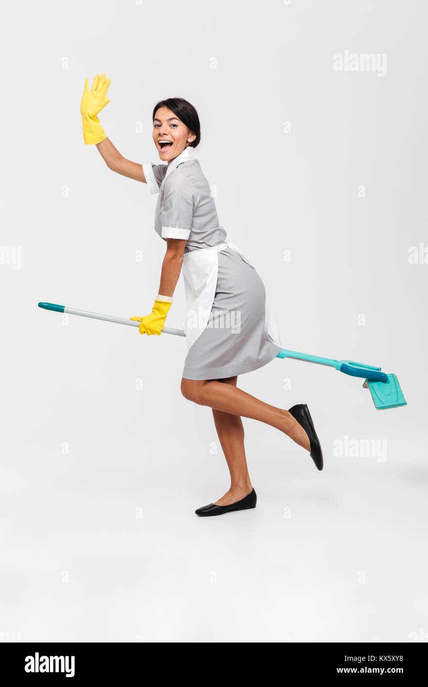 Full length photo of happy funny girl in uniform riding mop as a witch, isolated over white background Stock Photo