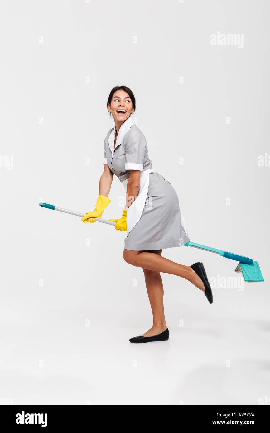 Full length photo of ridiculous young brunette woman in uniform and rubber gloves riding mop as witch, looking aside, isolated over white background Stock Photo