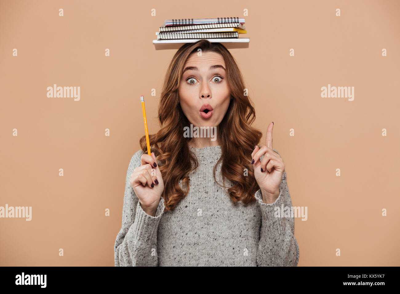 Close-up portrait of surprised female student in gray knitted sweater holding notebooks on her head and pointing with finger upward, isolated over bei Stock Photo