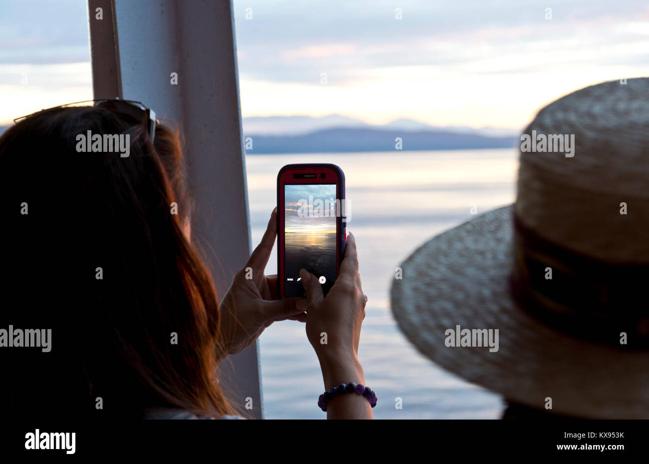 Woman taking a photograph with her cell phone of the sunset on the water.  On a BC Ferry on the Strait of Georgia. Stock Photo