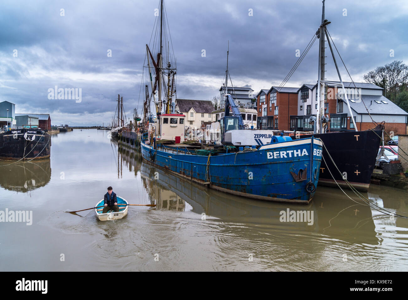 A man rowing a small boat past MV 'Bertha' and other ships at Anchorage Hill, Maldon, Essex, England, UK Stock Photo