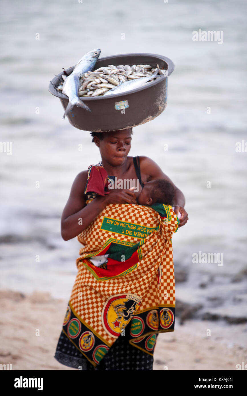 Mother breastfeeding baby while carrying bowl of fish on top of head, Inhambane, Mozambique Stock Photo