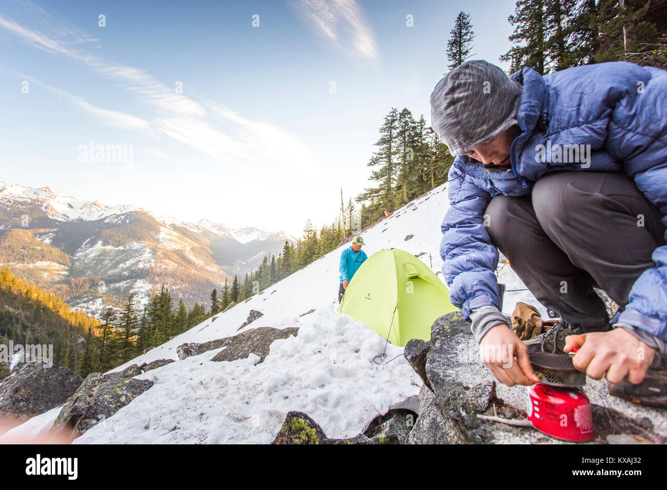 Woman crouching on snowy slope and lighting portable stove in front of man pitching tent, Leavenworth, Washington, USA Stock Photo