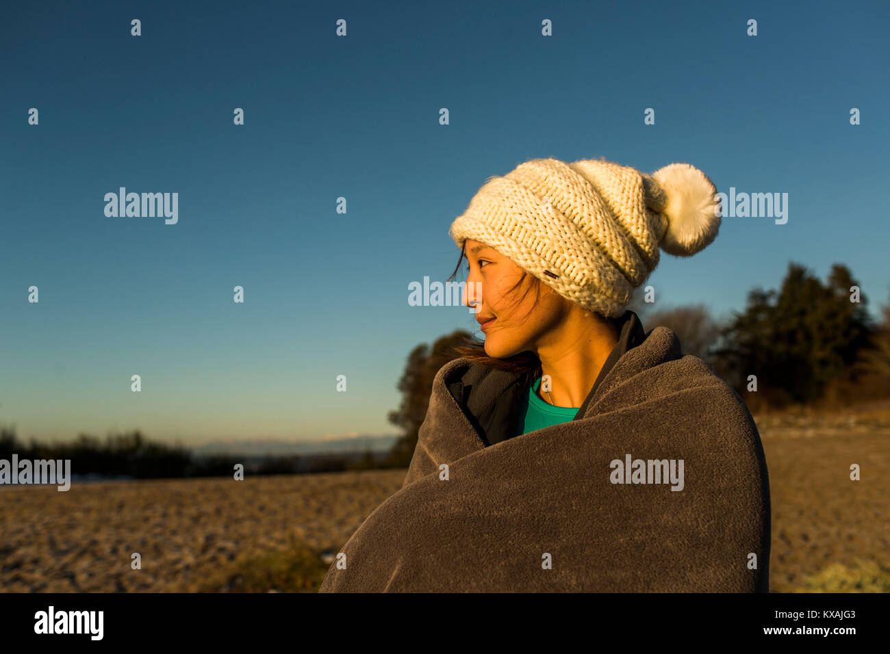 Young woman sitting in knit hat and wrapped in blanket while resting after running in Discovery Park, Seattle, Washington Sate, USA Stock Photo