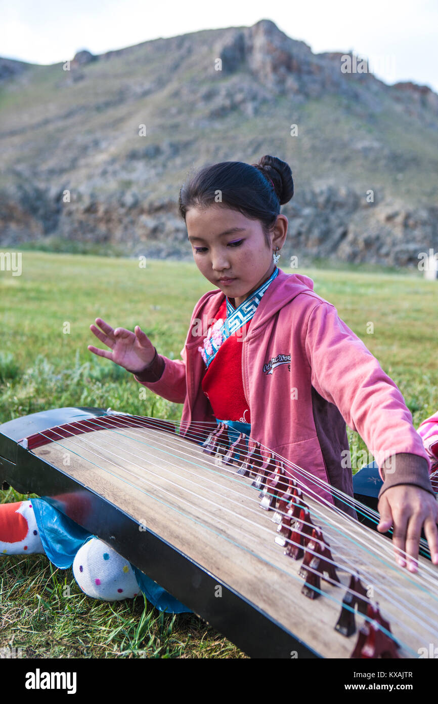 Young girl playing Yatga - Yatuga (string instrument) at Naadam Festival. Mongolia. The yatga is a half-tube zither with a movable bridge. It is constructed as a box with a convex surface and an end bent towards the ground. The strings are plucked and the sound is very smooth. The instrument was considered to be sacrosanct and playing it was a rite, bound to taboos. The instrument was mainly used at court and in monasteries, since strings symbolized the twelve levels of the palace hierarchy. Stock Photo