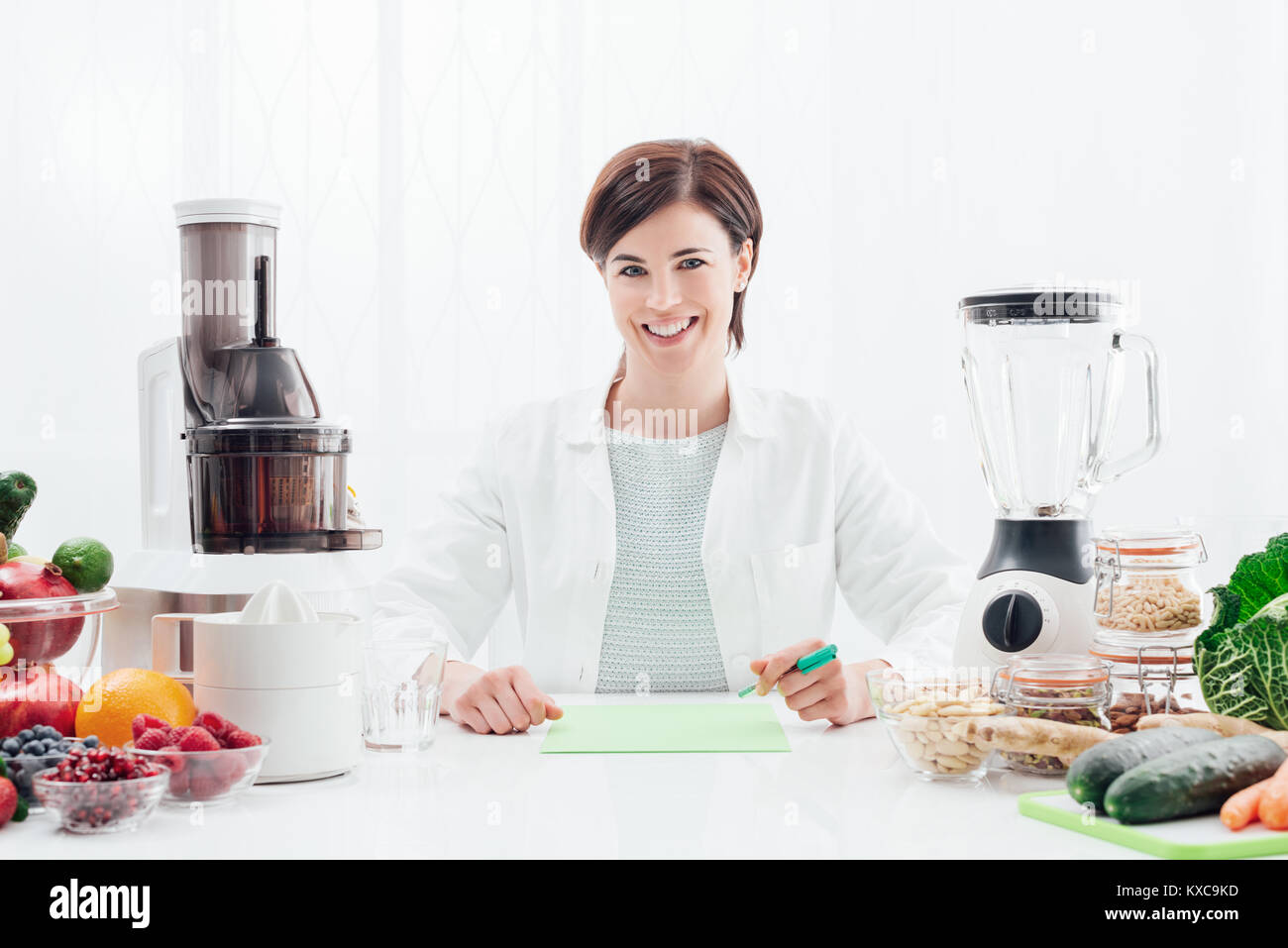 Smiling professional nutritionist holding an apple, she has healthy fruits, vegetables and juicers on her desk; healthy diet and wellness concept Stock Photo
