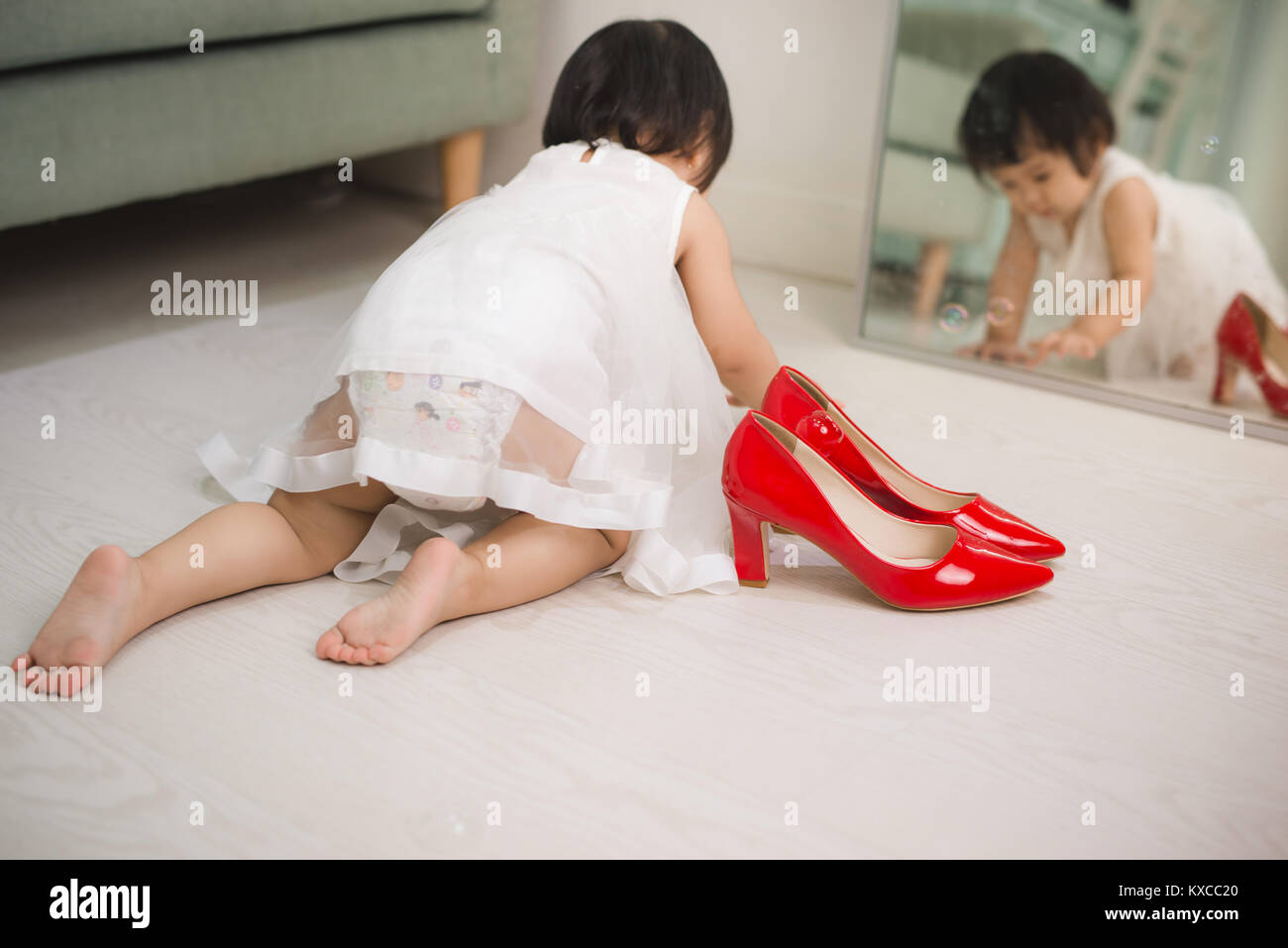 Cute funny little baby girl walks at home trying mom's red high heel shoes Stock Photo