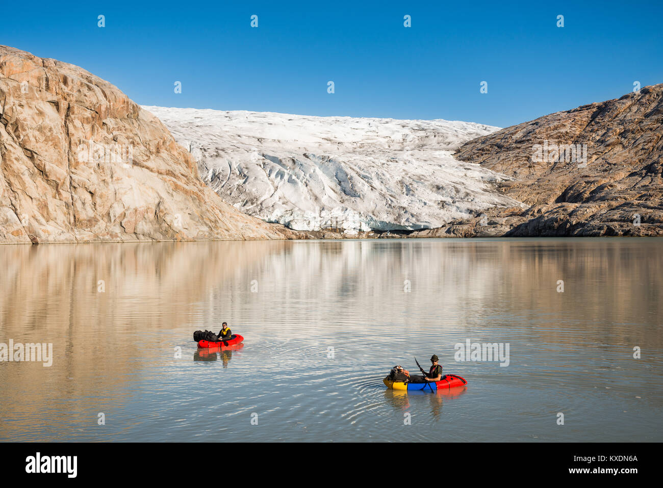 Two persons with pack rafts on fjord, behind glacier and rocks, blue sky, Greenland Stock Photo