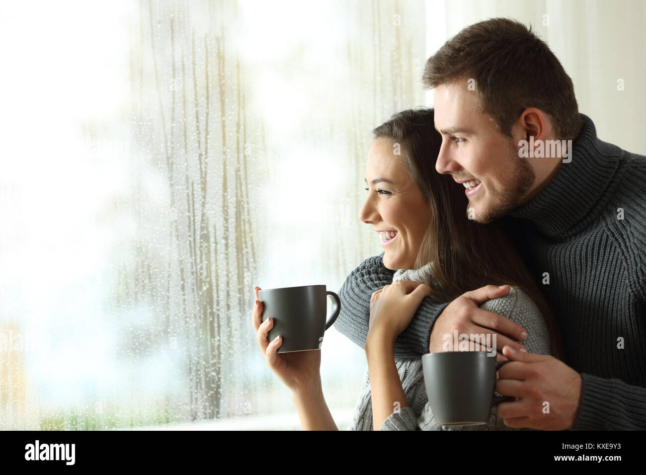 Side view portrait of a happy couple holding coffee mugs looking outside through a window a rainy day of winter at home Stock Photo