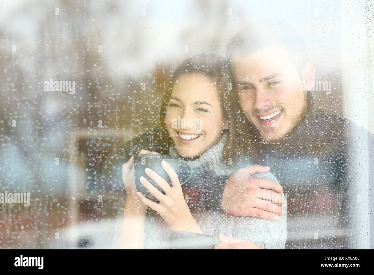 Front view portrait of a positive couple looking outdoors through a window in a rainy day of winter at home Stock Photo