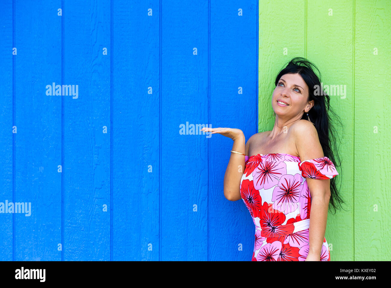 Young woman looking at her empty extended hand ready for your product placement as she poses against a wooden wall in tropical blue and green with cop Stock Photo
