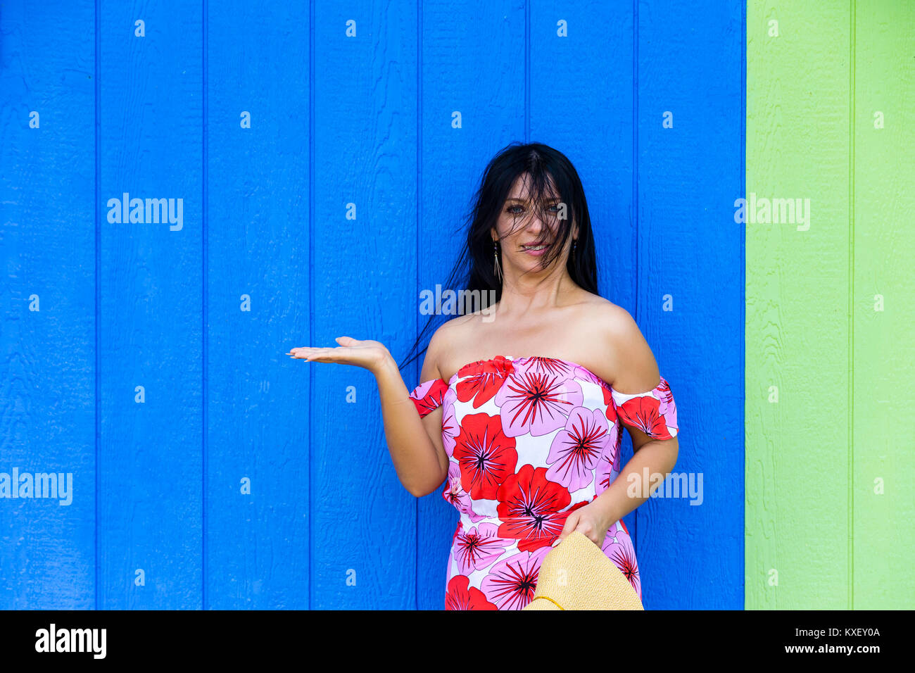 Pretty woman in a red floral summer dress standing against green and blue wooden wall with copy space holding out her empty hand for product placement Stock Photo