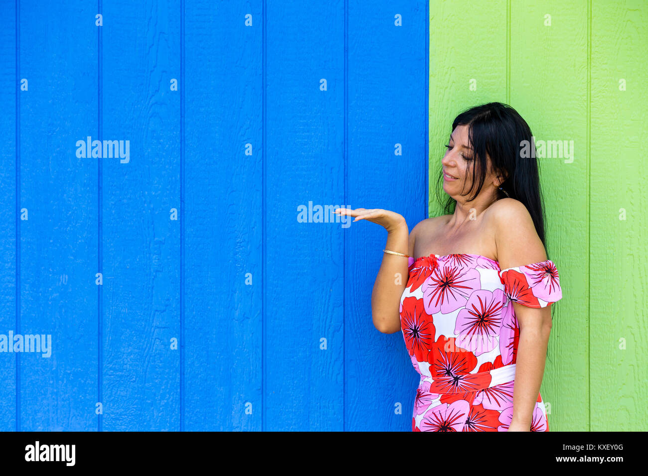Young woman looking at her empty hand extended for product placement as she stands in a red floral summer dress in front of a blue and green timber wa Stock Photo