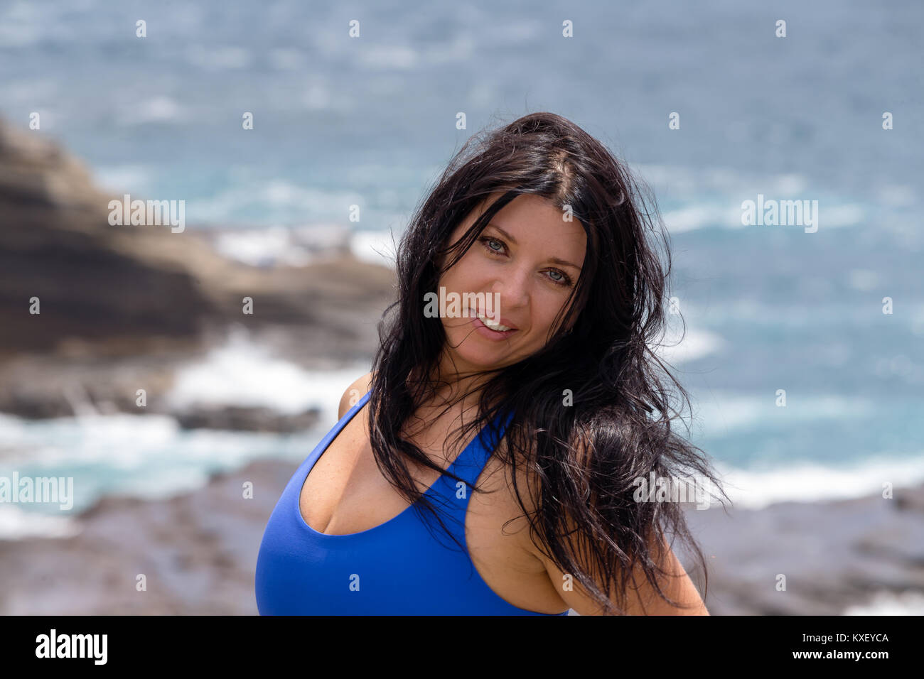Attractive windblown natural young woman with long tousled hair on the rocks at Heavens Point, Oahu, Hawaii smiling at camera Stock Photo