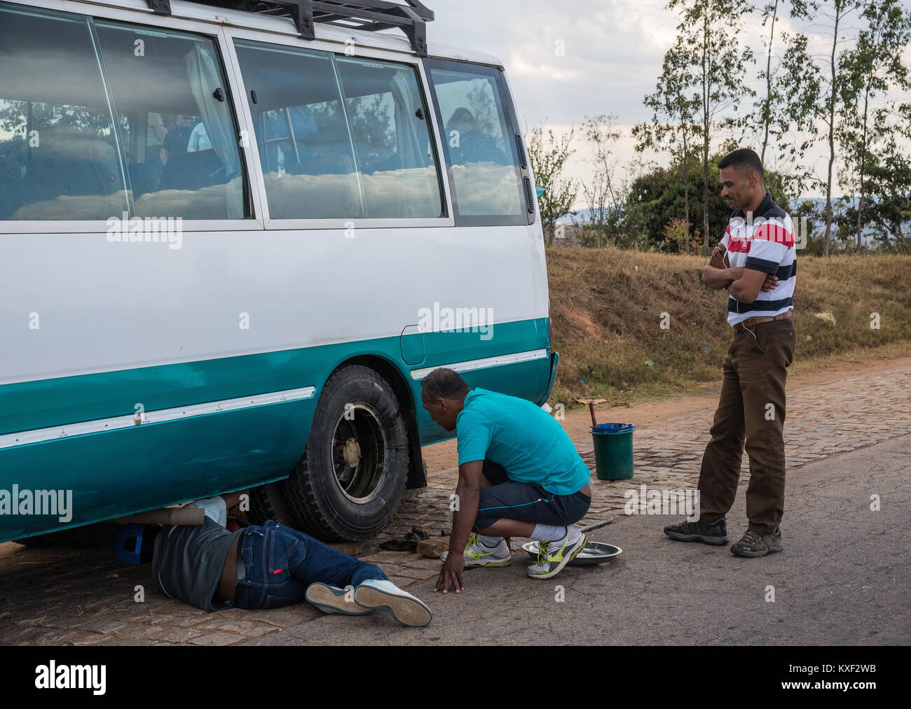 A man is under a minibus, trying to fix a flat tire, on the side of road. Madagascar, Africa. Stock Photo