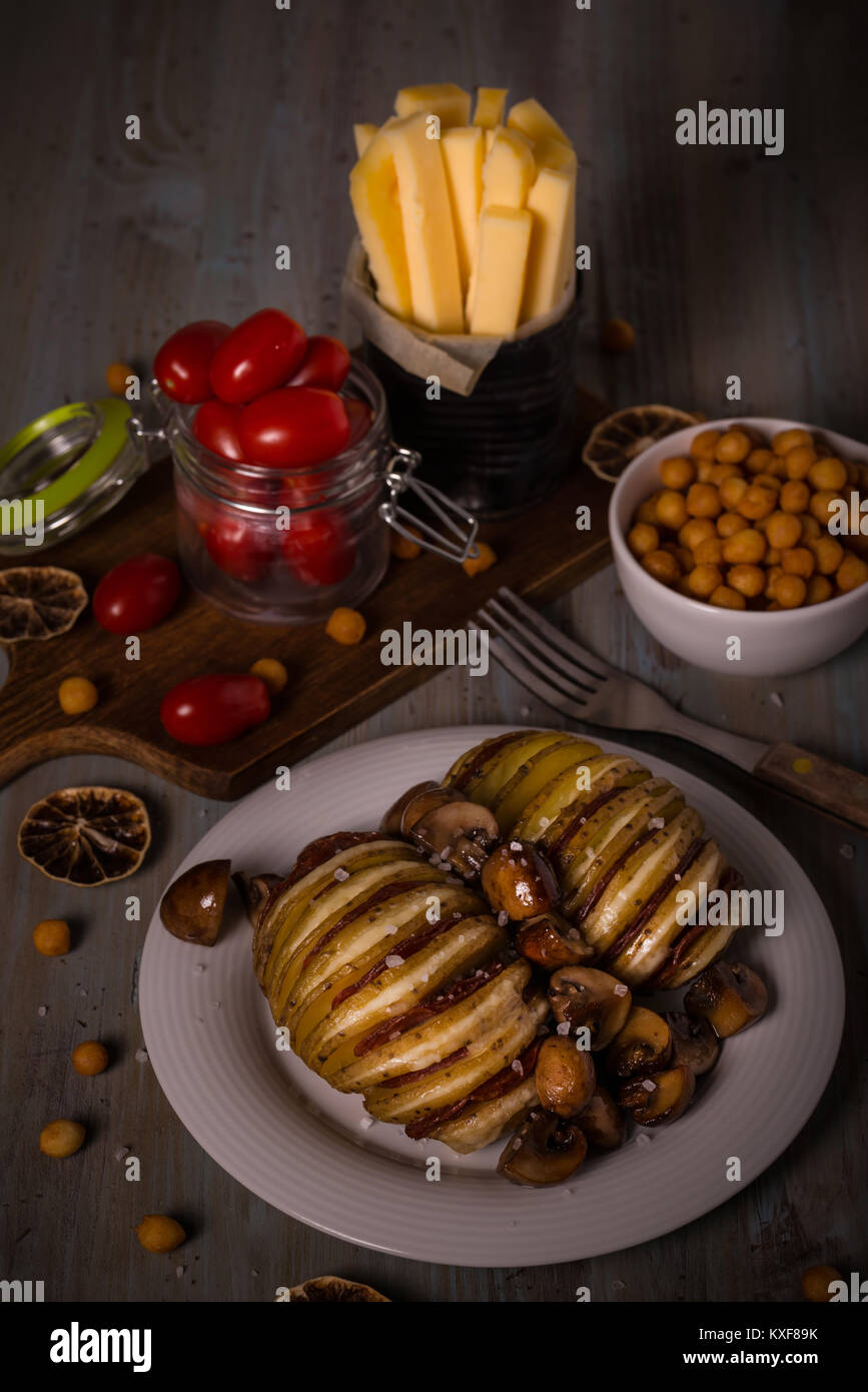 Vertical photo of two roasted and sliced full potatoes filled by gouda cheese and salami on white plate with fried brown mushrooms. Tomatoes, fried pe Stock Photo