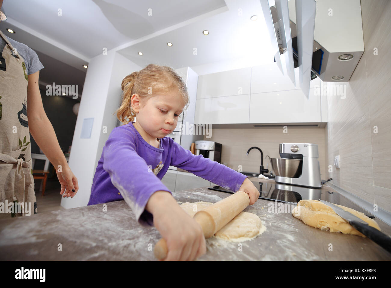 Little daughter helps mom to bake a cake. Toddler girl uses rolling pine in the kitchen. Stock Photo