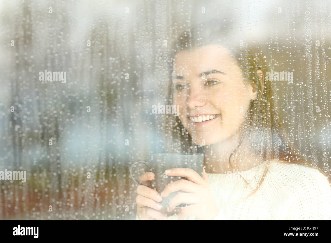 Positive happy teen looking through a window holding a coffee mug in a rainy day Stock Photo