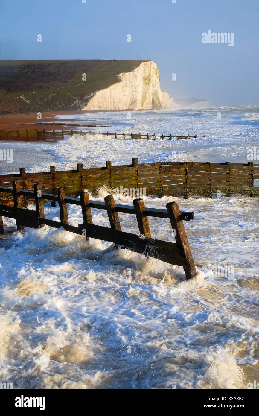 rough breaking white waves against wooden sea groynes in the forground looking accross sweeping pebble beach to white cliffs in background blue sky Stock Photo