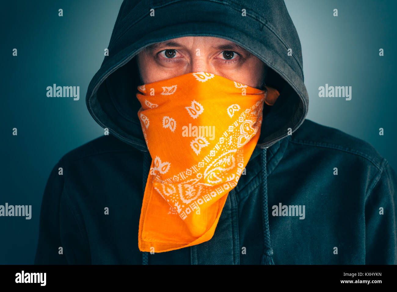 Portrait of masked criminal male person looking at camera. Adult man with hoodie and scarf over face as bandit or gang member. Stock Photo