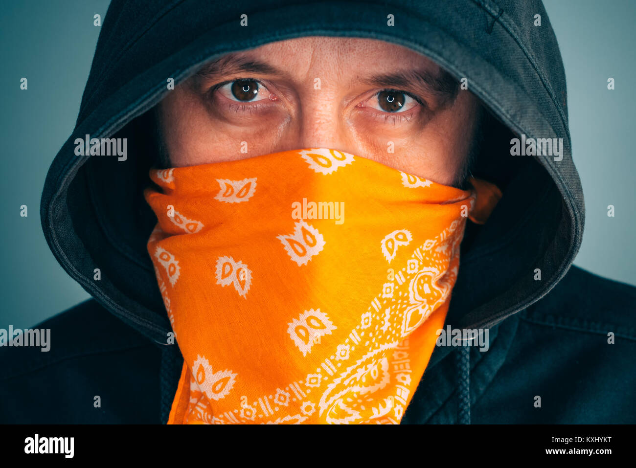 Portrait of masked criminal male person looking at camera. Adult man with hoodie and scarf over face as bandit or gang member, extreme close up Stock Photo