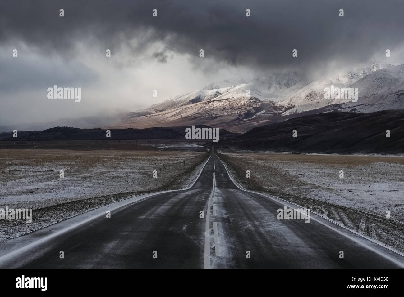 Road asphalt path on a winter snow desert wild mountain valley  at the background of the high peaks under a dramatic storm cloud Chuysky tract Altai M Stock Photo
