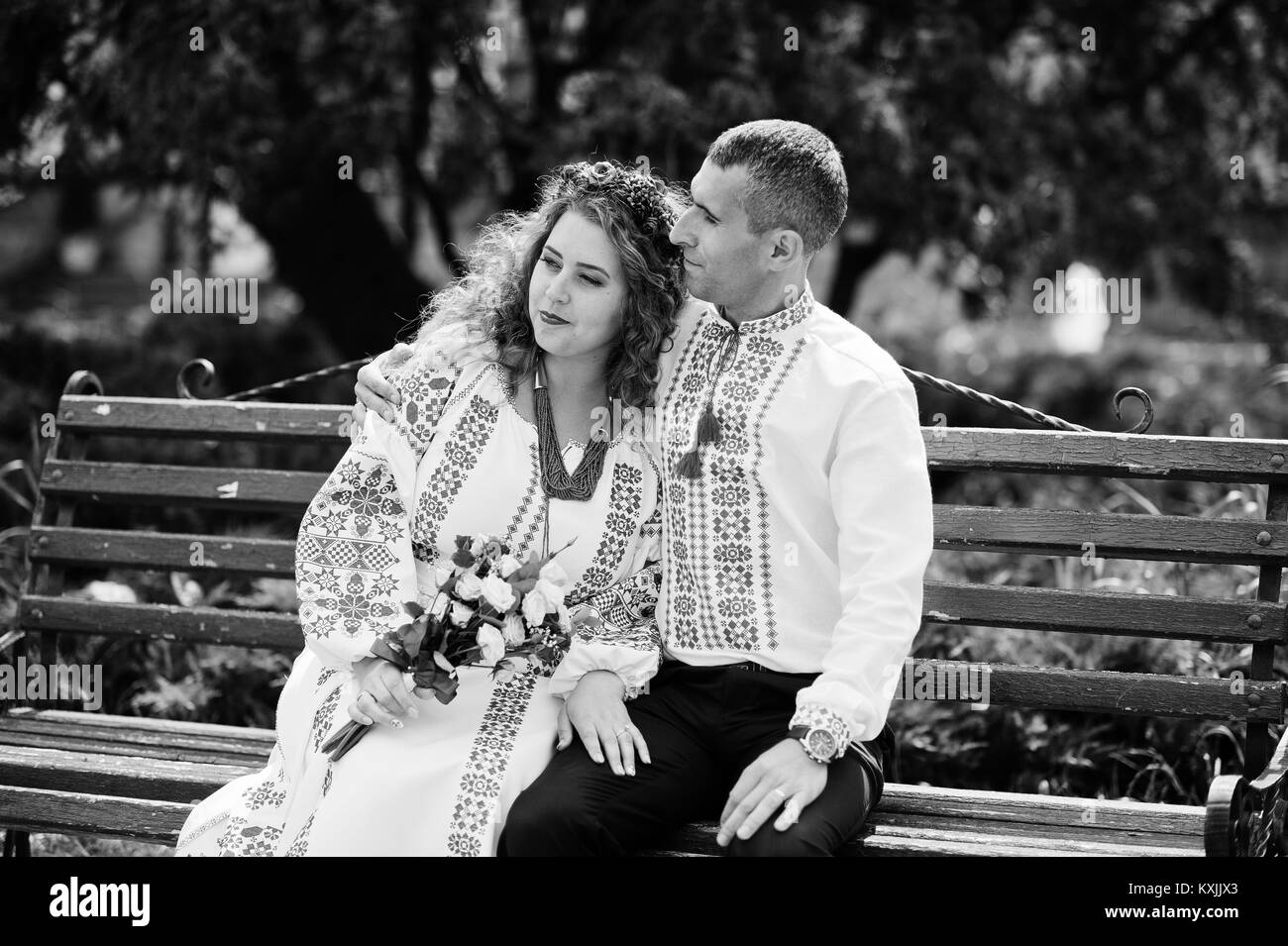 Cute wedding couple in ukrainian traditional clothes sitting on the bench in the town and spending time together. Black and white photo. Stock Photo