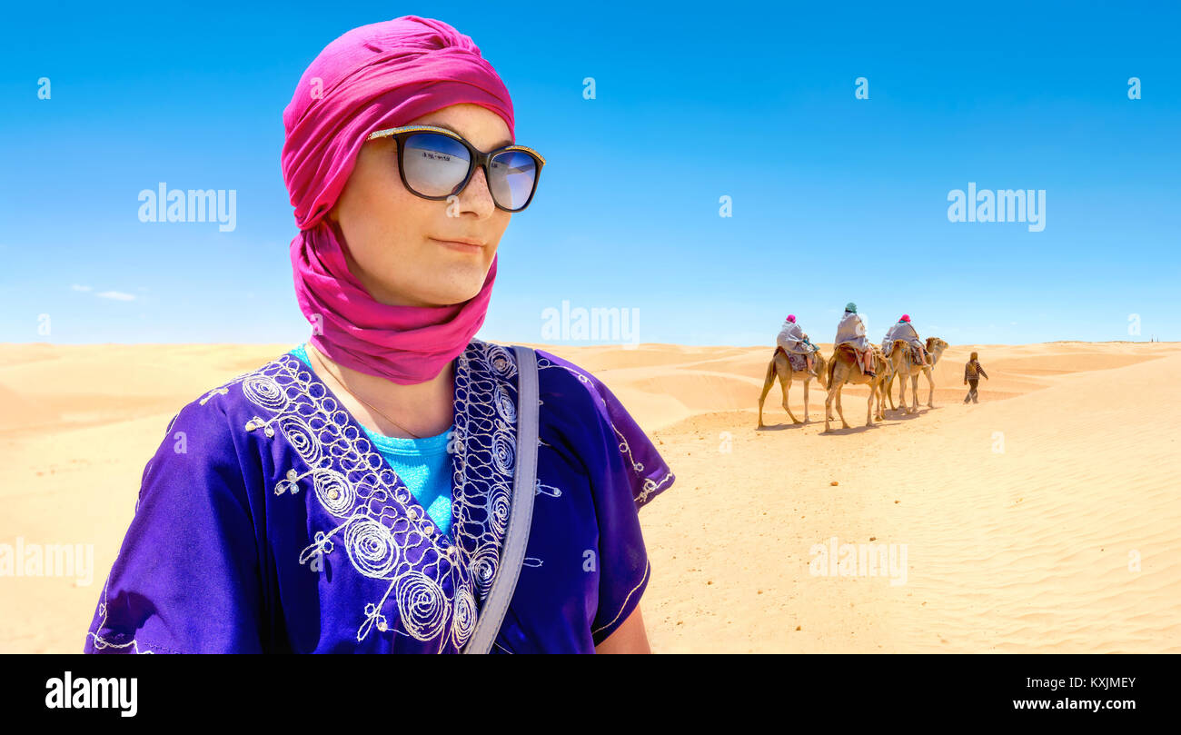 Portrait of beautiful woman in arabic traditional clothing against background of tourists riding on camels. Sahara desert, Tunisia, North Africa Stock Photo