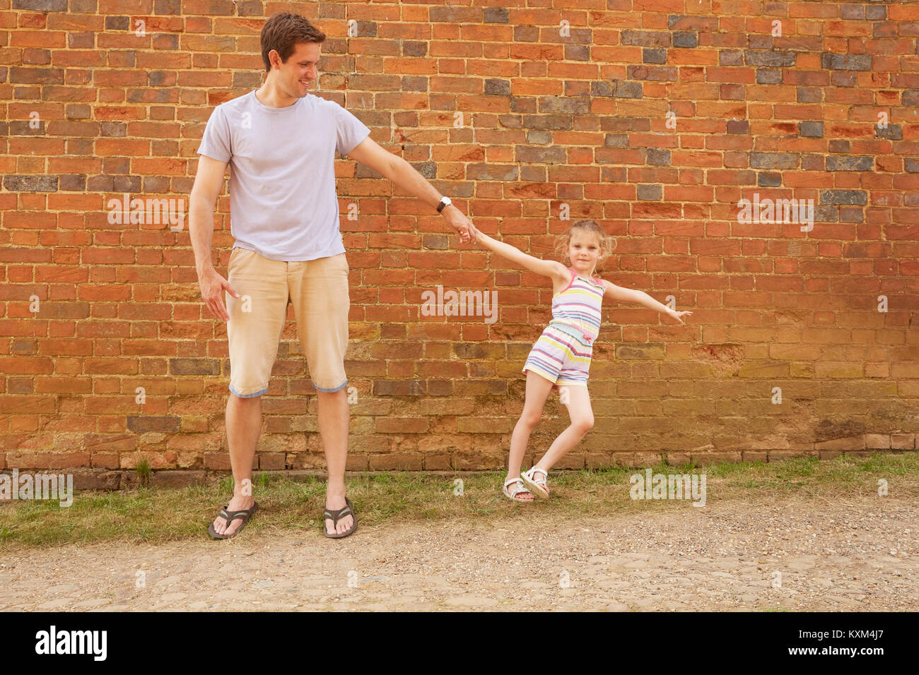 Girl leaning sideways holding father's hand by brick wall Stock Photo