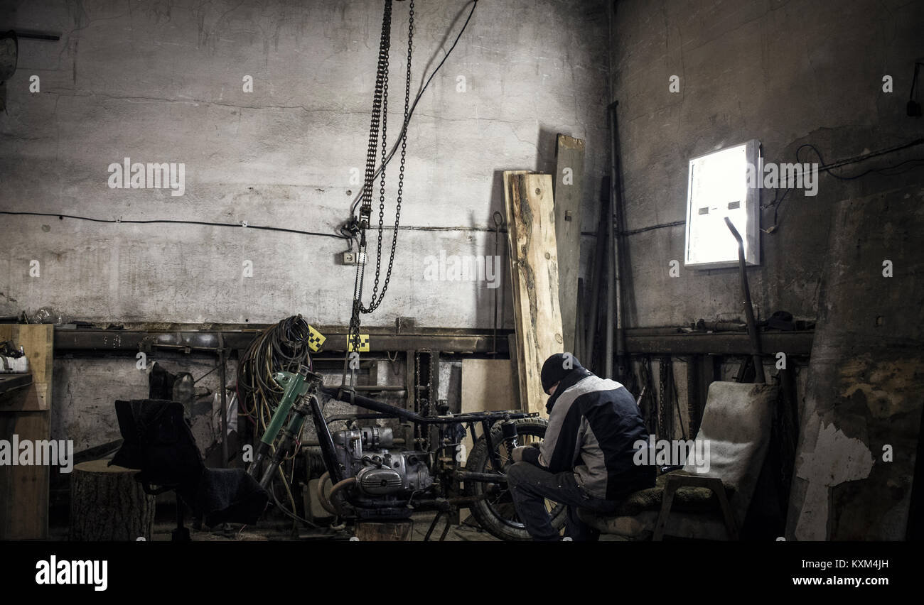 Mechanic sitting on armchair in workshop with dismantled vintage motorcycle Stock Photo