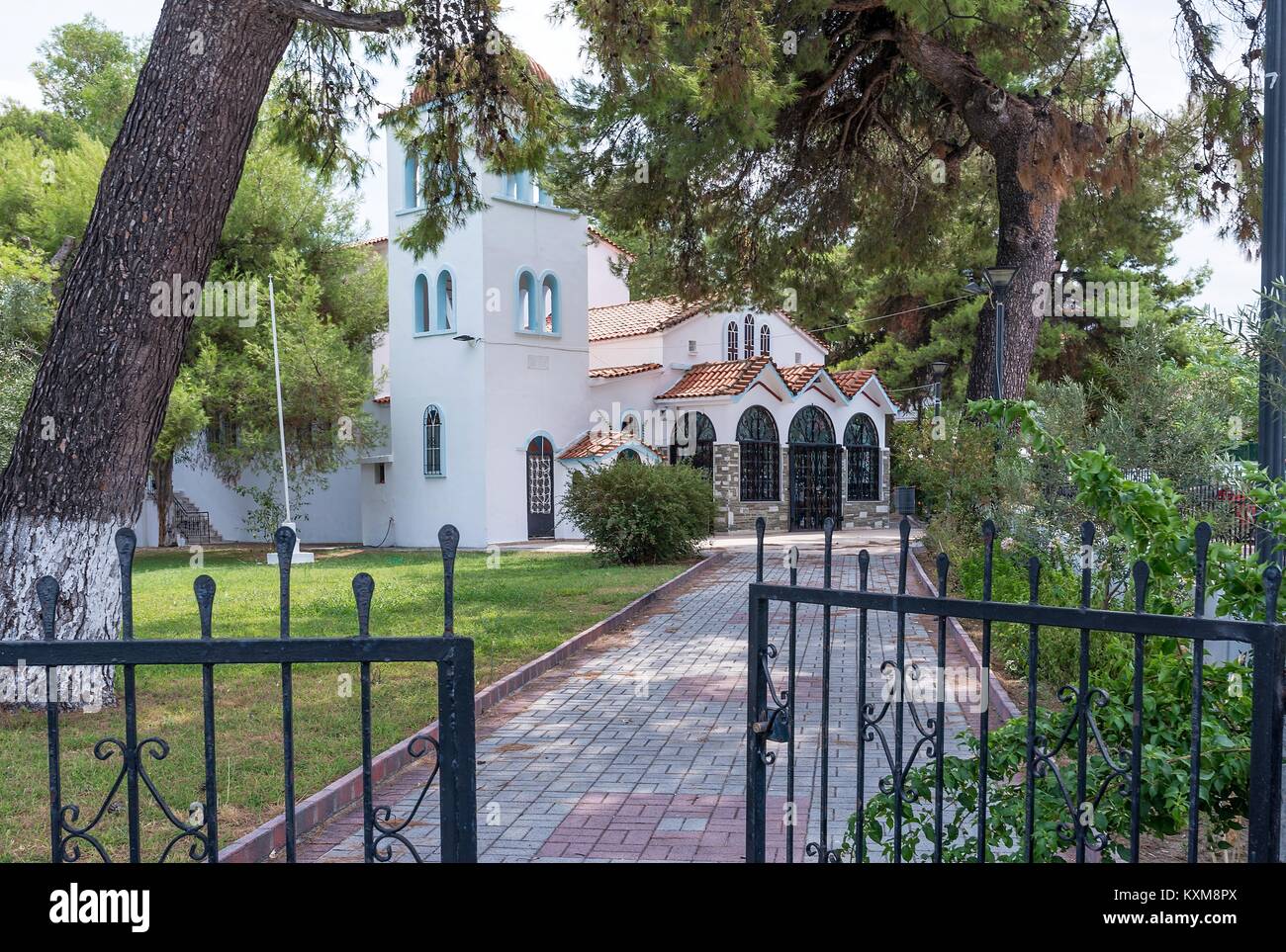 View of a Greek Orthodox Church where a paved path leads underneath an overarching tree to the church entrance. Stock Photo