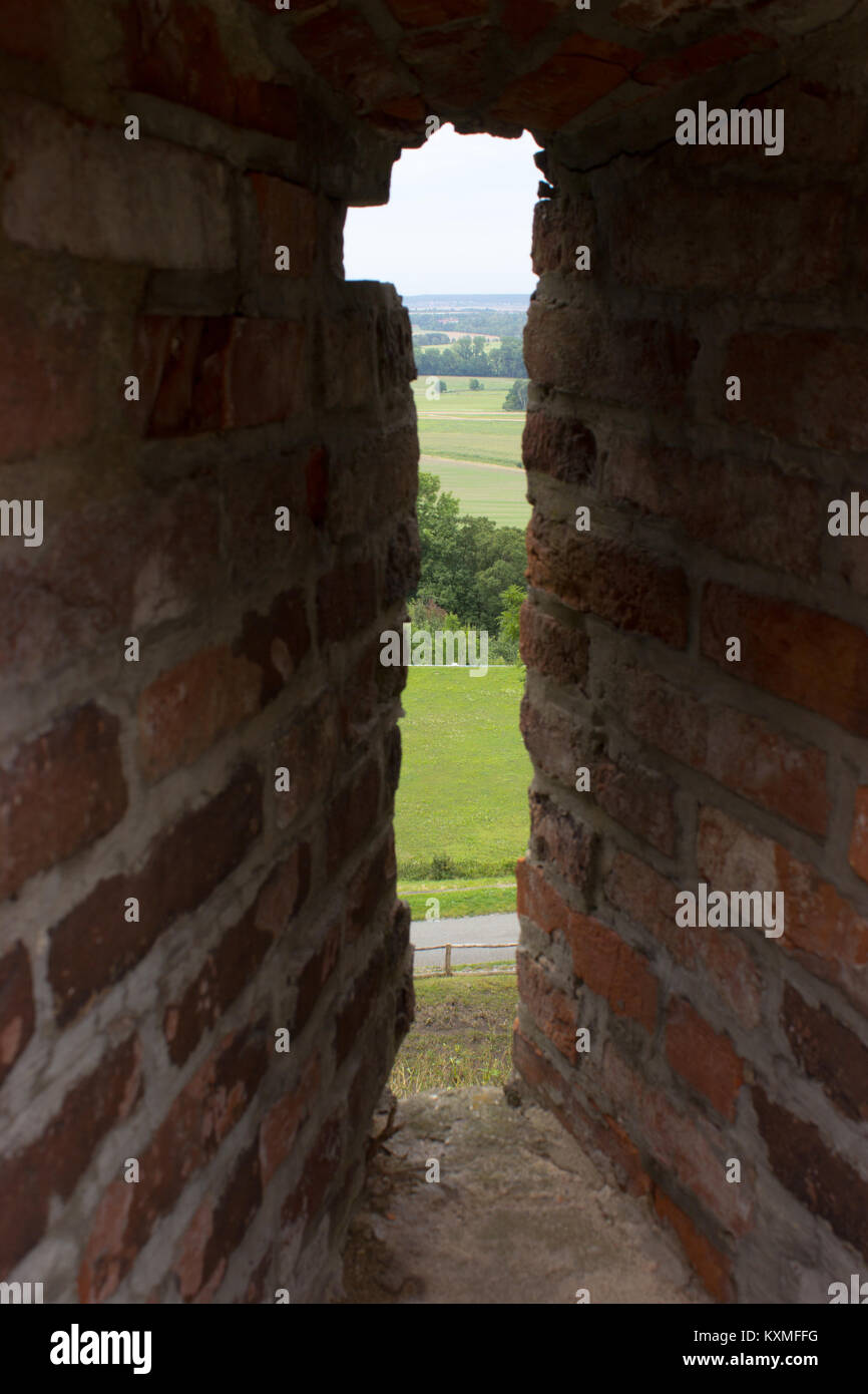 A view across the castle loophole on the landscape. In the foreground is a loophole from the bricks, in the background is a path, a meadow, a city. Stock Photo