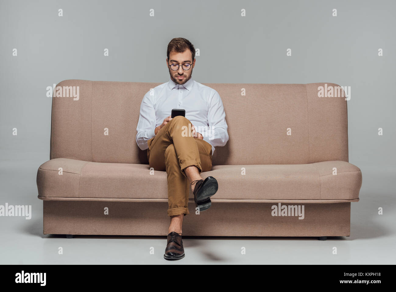 stylish man in eyeglasses sitting on couch and using smarttphone isolated on grey Stock Photo
