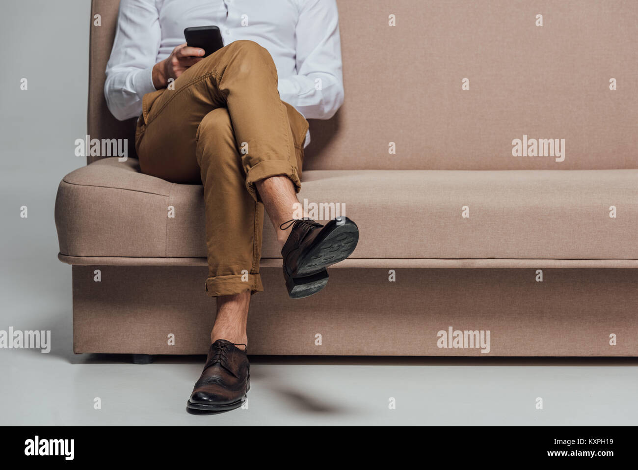 low section of young man using smartphone while sitting on sofa isolated on grey Stock Photo