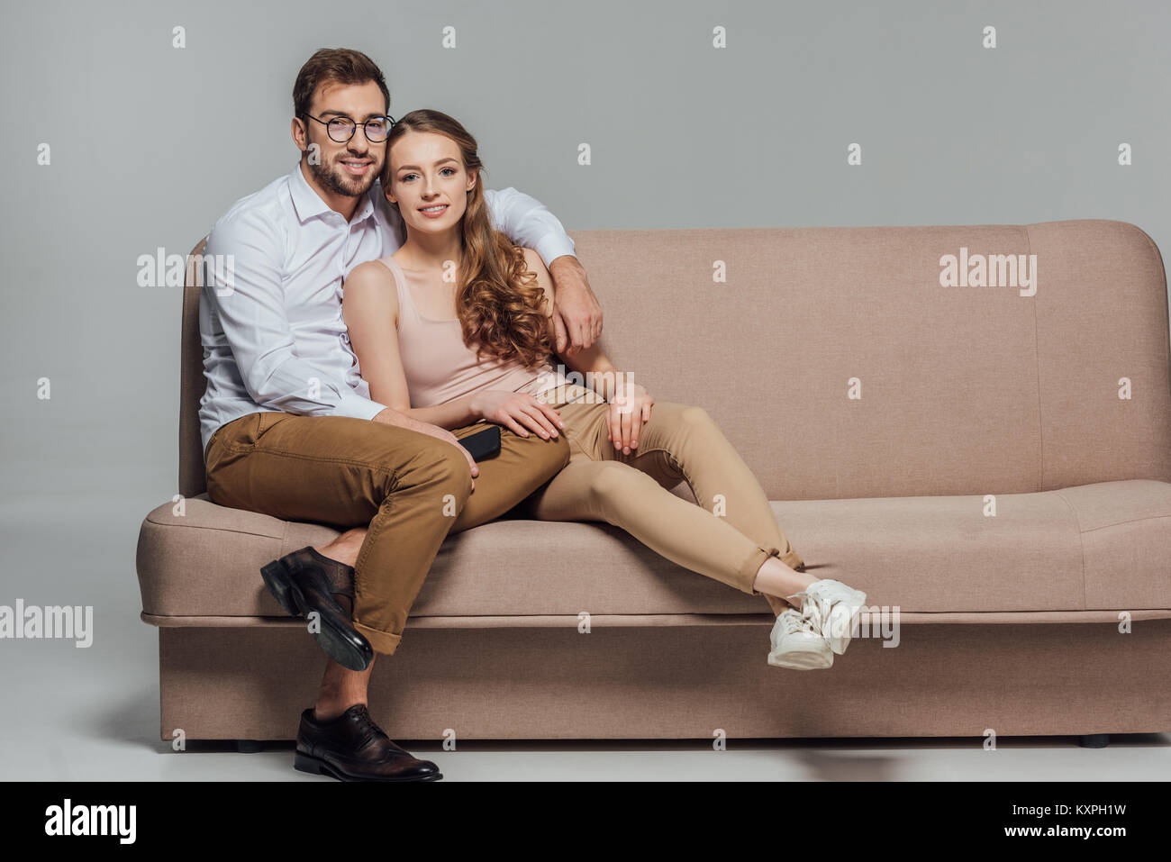 happy stylish young couple with smartphone smiling at camera while sitting together on sofa isolated on grey Stock Photo