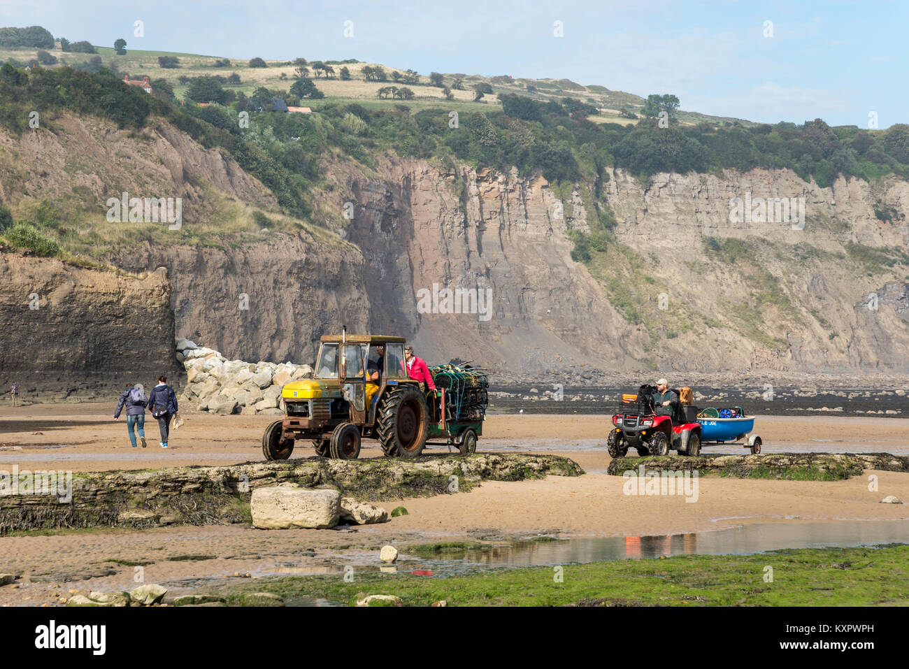 Tractor bringing in Lobster pots ahead of a quad bike and small boat on the beach at Robin Hood's Bay, North Yorkshire, England. Stock Photo