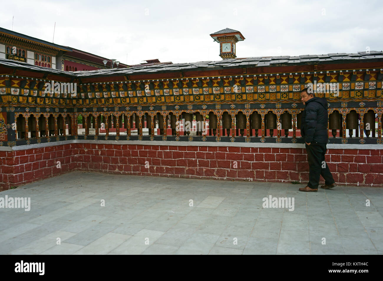 Man truning prayer wheels in downtown Thimphu. Above is the clock of the Clocktower, Bhutan Stock Photo