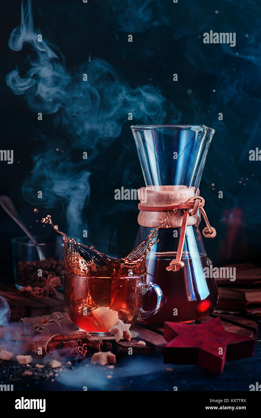 Glass coffee cup with a dynamic splash and rising steam. Hourglass-shaped coffeemaker with on a dark background with copy space. Action food photograp Stock Photo