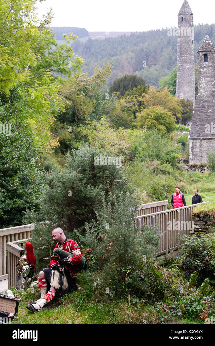 Man busking dressed in Scottish Soldier Outfit sitting by his armor and playing bagpipes at the entrance to the monastic site in Glendalough Valley Stock Photo