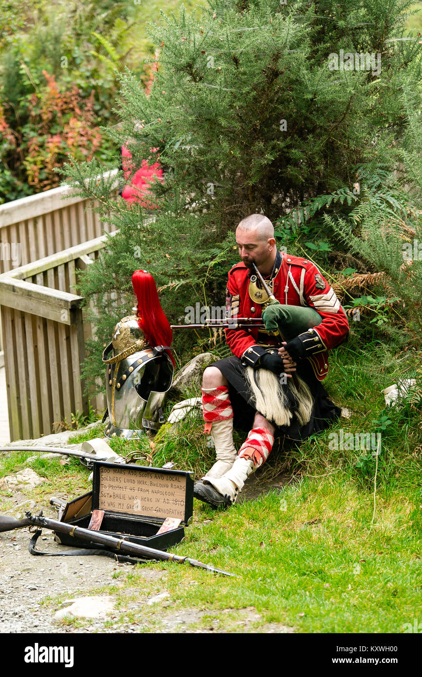 Man busking dressed in Scottish Soldier Outfit sitting by his armor and playing bagpipes at the entrance to the monastic site in Glendalough Valley Stock Photo