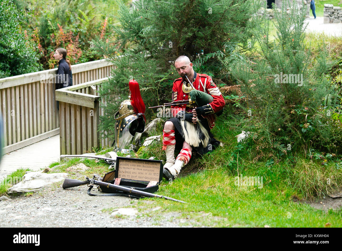 Man busking dressed in Scottish Soldier Outfit sitting by his armor and playing bagpipes at the entrance to the monastic site in Glendalough Valley Stock Photo